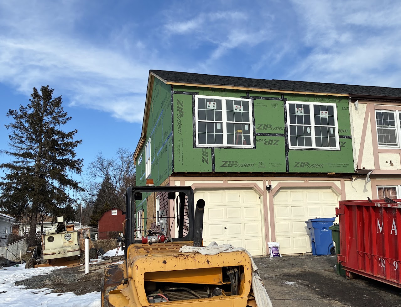 A two-storey addition in green structural sheathing with windows set, a skid steer in the foreground
