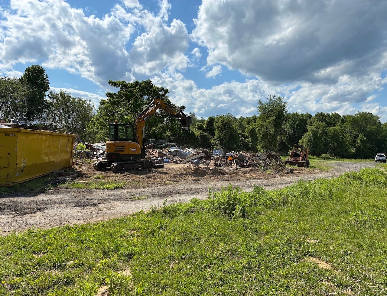 An excavator breaking up a cleared lot beside a yellow dumpster